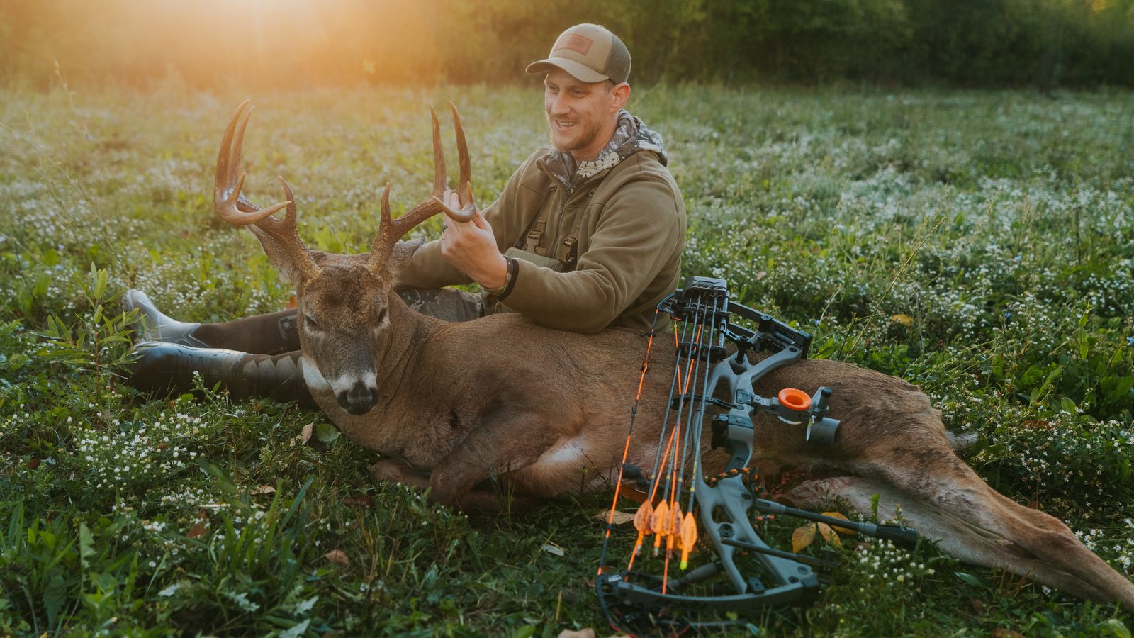 Bowhunter with his whitetail buck and compound bow in a field at sunrise