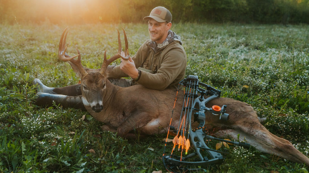 Bowhunter with his whitetail buck and compound bow in a field at sunrise